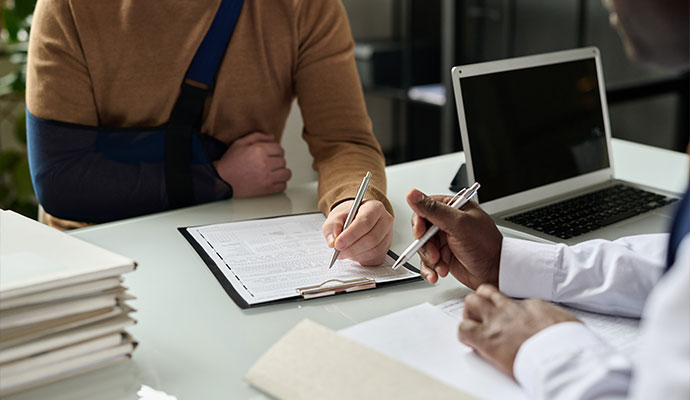 A person with their arm in a dark blue sling meeting with a professional at a desk A person with their arm in a dark blue sling meeting with a professional at a desk